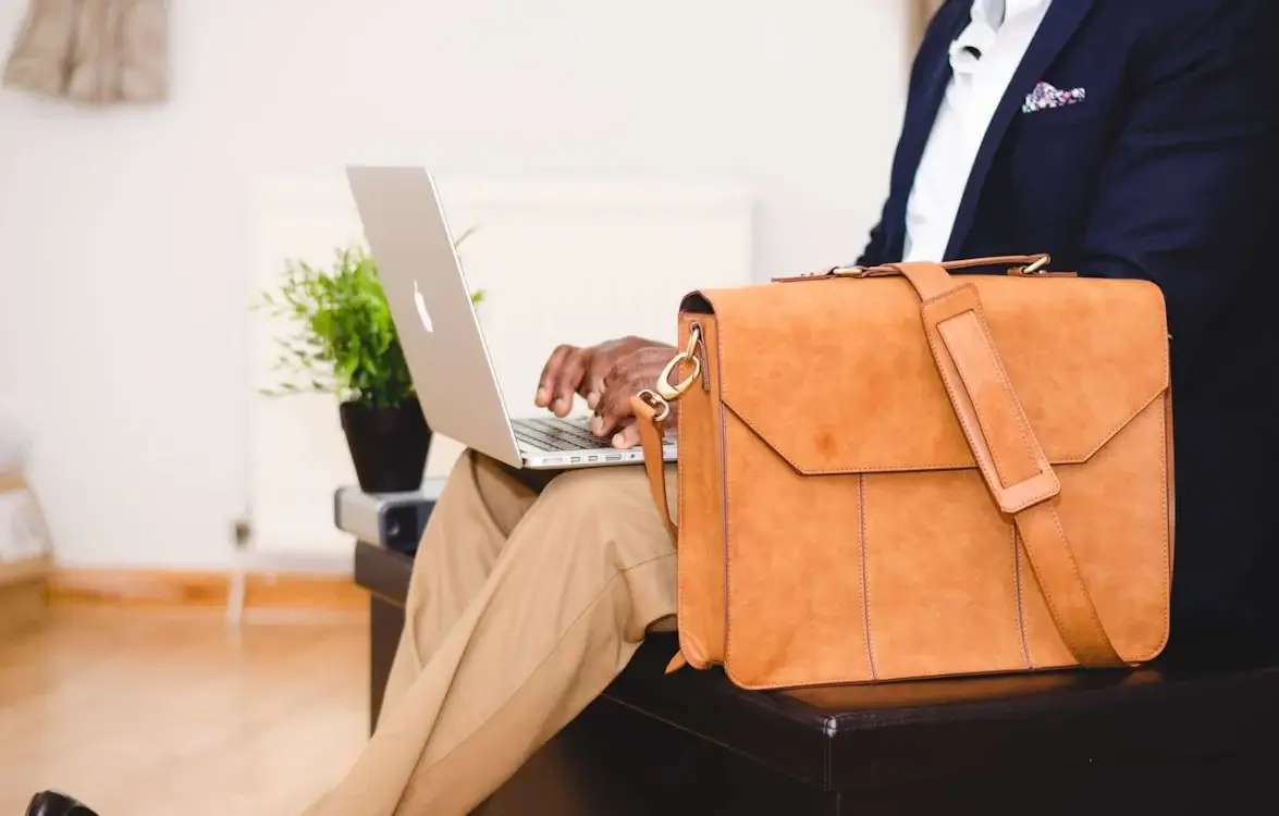 person wearing blue jacket, seated next to tan briefcase, working on laptop